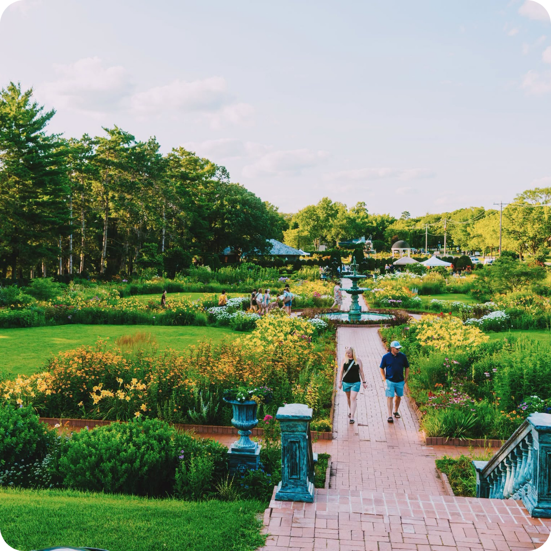 A couple walks along a brick pathway in a colorful garden filled with flowers and greenery, centered around a decorative fountain.